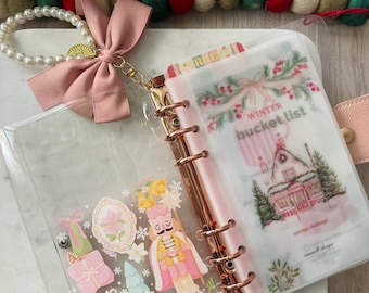 Könnte beinhalten: Ein transparenter A6-Kunststoffordner mit roségoldenen Ringen, verziert mit einem Nussknacker- und Weihnachtsgeschenkmotiv auf dem Einband. Eine rosa Schleife und ein Perlenanhänger sind oben befestigt. Innen zeigt eine halbtransparente Seite eine Liste.
