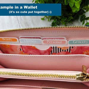 Puede incluir: Una cartera rosa con una cremallera dorada est&aacute; abierta para revelar cinco divisores etiquetados como "Belleza", "Ropa", "Personal", "Comer fuera" y "Comida".