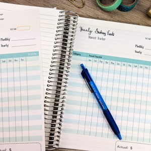 May include: A close-up of a blue spiral bound notebook with a light blue and white gridded page. The page is labeled "Yearly Saving Funds Deposit Tracker" and has columns for category, amount deposited, and months of the year. A blue pen is lying on the page.