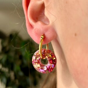 May include: Gold hoop earrings featuring a circular charm filled with pink, red, and gold glitter. The earrings are shown on an ear. The charm has a donut shape.