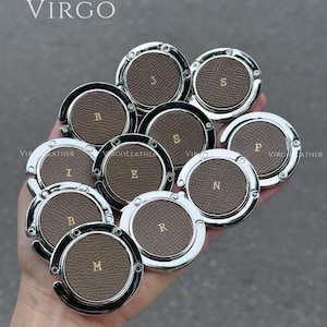 Può includere: Una collezione di appendini per borse in pelle marrone con bordi in metallo argentato, ognuno con una lettera dorata. La parola "VIRGO" è stampata in alto. Gli appendini rotondi hanno un gancio per appendere le borse.