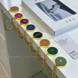 Op de afbeelding: Een collectie van zeven goudkleurige metalen tashaken met ronde lederen inzetstukken in diverse kleuren, waaronder lichtbruin, rood, bordeaux, blauw, zwart, mosterdgeel en groen, elk voorzien van gepersonaliseerde, goudkleurige initialen of namen.