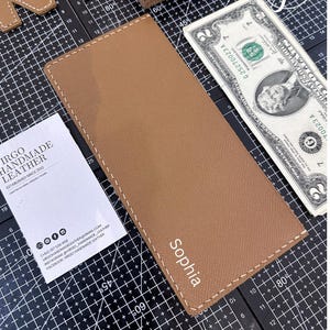 May include: A tan leather wallet with white stitching and the name "Sophia" printed on the front. The wallet is rectangular and lies on a black surface with a two-dollar note and a business card.