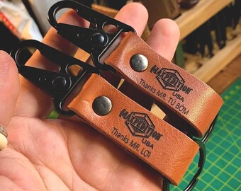 May include: Two tan leather keychains featuring black metal carabiner clips and black metal key rings. Each leather strap is secured with a silver-tone rivet and embossed with the Maxpedition USA logo, along with the text Thanks MR. TU BOM or Thanks MR. LOI.