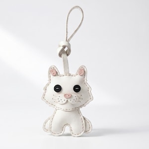 May include: A white leather cat-shaped keychain featuring two black button eyes, a small pink stitched nose, and pink inner ear details. The cat figure is attached to a matching white leather loop strap with visible stitching along the edges.