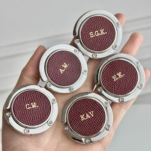 Puede incluir: Cinco colgadores de bolso plateados con inserciones de cuero burdeos. Cada colgador presenta iniciales doradas diferentes: S.G.K., A.W., E.K., C.W. y K.A.V. Los colgadores tienen un diseño circular con un gancho plegable.