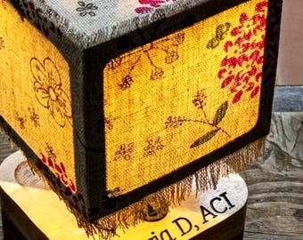 May include: A cube-shaped lamp with a floral design on the top and sides. The lamp has a wooden base with the text "Maria D, ACI" engraved on it. The lamp's sides are illuminated, showcasing the floral pattern.