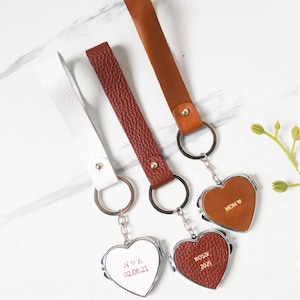 Puede incluir: Llaveros de cuero hechos a mano en blanco, burdeos y marrón. Cada llavero presenta una correa de cuero y un medallón en forma de corazón con texto grabado. El texto incluye nombres, fechas y la palabra "MOM". Los llaveros están sujetos a llaveros plateados.