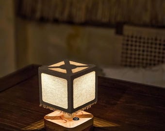 May include: A cube-shaped table lamp with a wooden base and a fabric shade. The shade is dark with a lighter fabric panel on each side. The lamp is on a dark wooden table, casting a warm glow.