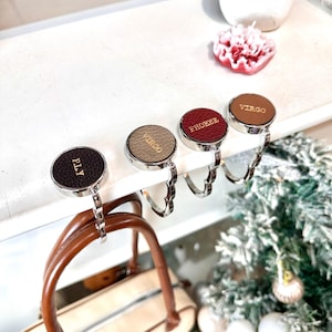 May include: Four silver handbag hooks with leather covers. The covers are in different colors and have names written on them: "PLY", "Virgo", "Phoebe", and "Virgo".