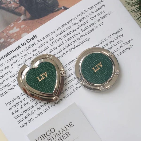 Könnte beinhalten: Zwei silberfarbene Handtaschenhaken mit grünen Ledereinsätzen. Einer ist herzförmig, der andere rund, beide mit "LIV" in goldfarbenen Buchstaben. Die Haken liegen auf einer weißen Oberfläche mit gedrucktem Text und einem Logo.