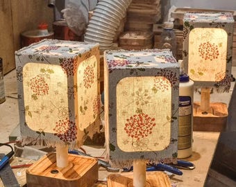 May include: Three handmade table lamps with square wooden bases and floral patterned shades. The shades are a light gray with red and white floral designs. The lamps have a warm, inviting glow, perfect for a bedroom or living room.