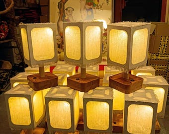 May include: A collection of square lamps with a warm, inviting glow. Each lamp features a wooden base and a square shade with a textured, off-white fabric. The lamps are arranged in a group, suggesting they are for sale or display.