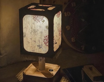 May include: A square-shaped table lamp with a floral patterned shade. The shade is black with a linen-like fabric that has a floral design in red and white. The base is made of wood. The lamp is turned on, illuminating the floral pattern.