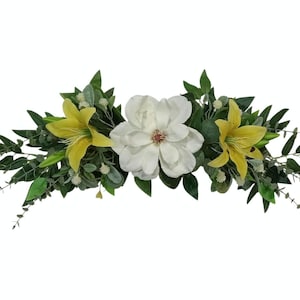 May include: A floral arrangement featuring a large white magnolia flower flanked by two yellow lilies. Green leaves and small white flowers fill the spaces between the blooms. The arrangement is designed to be a centerpiece.