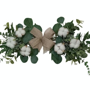 May include: A decorative floral arrangement featuring white cotton bolls, green eucalyptus leaves, and small white flowers. A burlap bow is centered in the design. This arrangement is suitable for home decor.