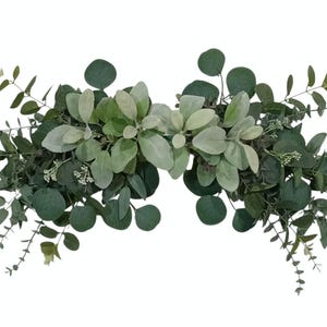 May include: A decorative floral arrangement featuring a variety of green foliage. The arrangement includes round eucalyptus leaves, small white flowers, and soft, light green lamb's ear leaves. The design is symmetrical and ideal for home decor.
