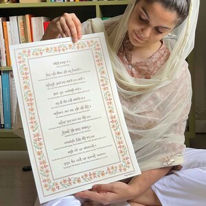 May include: A framed print with text in a foreign script, featuring a floral border. The print is held by a person. The border is decorated with peach and green floral elements.