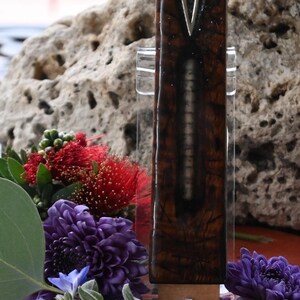 May include: A decorative wooden mezuzah case with a silver-colored letter "V" on the front. The case is displayed with purple and red flowers, green leaves, and a large rock in the background. The mezuzah case is approximately 6 inches tall.