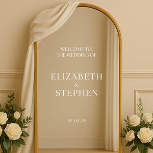 May include: A gold arched mirror with the text "WELCOME TO THE WEDDING OF ELIZABETH & STEPHEN 10.08.25". A cream-colored drape hangs over the top. Two bouquets of white roses and greenery are on either side of the mirror.