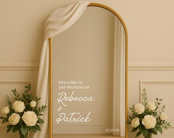 May include: A gold arched mirror with a cream-colored drape and the text "WELCOME TO THE WEDDING OF Rebecca & Patrick 12.10.25". Two bouquets of cream-colored roses flank the mirror. The background is a light beige color.