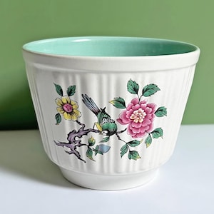 May include: White ceramic plant pot with a ribbed exterior and a pale green interior. The pot features a colourful floral and bird design with pink, yellow, green, and blue accents. A decorative piece for indoor plants.