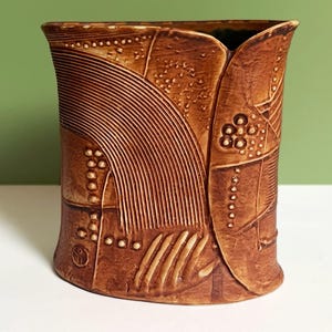 Könnte beinhalten: Eine braune Keramikvase mit strukturierter Oberfläche, die geschnitzte geometrische Muster wie parallele Linien, kleine erhabene Punkte und segmentierte Felder aufweist. Das Gefäß hat eine organische, leicht abgeflachte zylindrische Form.