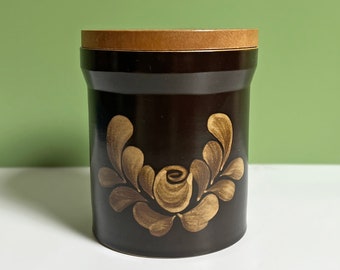 May include: A brown ceramic jar with a wooden lid and a gold floral design. The jar has a simple, elegant design and would be a great addition to any kitchen or bathroom.