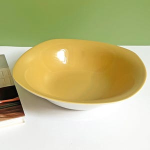 Könnte beinhalten: Eine große, oval geformte Keramikschale mit gelbem Innenraum und weißem Außenbereich. Die Schale steht neben einem Buch mit dem Titel "Mid-Century Modern at Home". Der Hintergrund ist einfarbig grün.