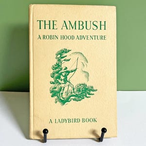 May include: Vintage book titled "The Ambush: A Robin Hood Adventure" by Ladybird Books. The cover is a light beige with green text and a green illustration of Robin Hood with a bow and arrow. The book is propped up on a black stand.