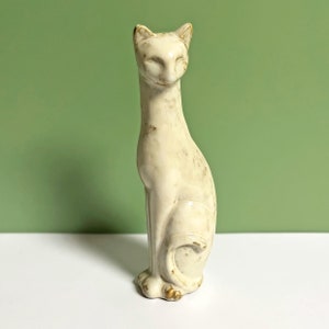 Może przedstawiać: Kremowa ceramiczna figurka kota w pozycji siedzącej. Kot ma długą szyję, zamknięte oczy i spiczaste uszy. Figurka ma brązowe akcenty i stoi na białej powierzchni z zielonym tłem.