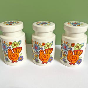 Może przedstawiać: Trzy białe ceramiczne pojemniki z pasującymi pokrywkami, każdy z kolorowym nadrukiem graficznym przedstawiającym pomarańczowego ptaka w otoczeniu stylizowanych niebieskich, żółtych i fioletowych kwiatów. Cylindryczne naczynia mają gładkie wykończenie.