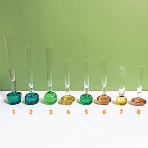 May include: Eight glass bud vases with different coloured bases. The bases are green, yellow, and orange. The vases are arranged in a row from left to right, with the largest vase on the left and the smallest vase on the right.
