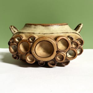 May include: A ceramic vessel with a wide, flared rim and two small handles on the sides. The exterior features a textured, cream-coloured glaze with numerous raised, circular, brown-rimmed protrusions of varying sizes across the lower body.