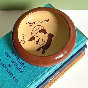May include: A small, round, ceramic bowl with a glossy brown rim and a cream-colored interior. The center features a dark brown hand-painted illustration of a person wearing a cap, with the handwritten text "Bill Brewer" inscribed above.