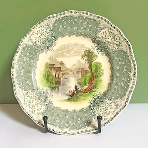 May include: A round ceramic plate with a scalloped edge featuring a central transferware scene of a river landscape with classical ruins, trees, and figures. The border is decorated with a green and white floral and stippled pattern.