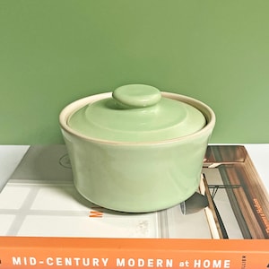 May include: A pale green ceramic covered dish with a lid. The dish is sitting on a book titled "Mid-Century Modern at Home".