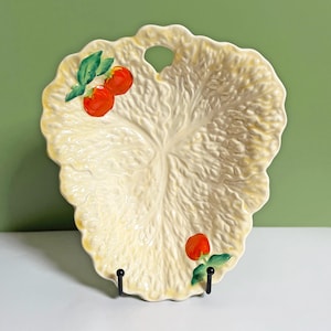 May include: Cream-colored, leaf-shaped ceramic serving platter with a scalloped edge and a handle. The platter is decorated with red tomato and green leaf accents. The platter is displayed on a black metal stand.