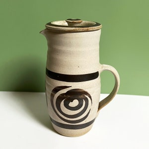 Puede incluir: Una jarra de cerámica blanca con una tapa marrón y un diseño de espiral negro. La jarra tiene un asa y un pico.