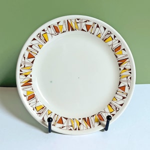 Op de afbeelding: Een witte keramische plaat met een geometrisch patroon in bruin-, geel- en oranje tinten rond de rand. De plaat is rond en lijkt vintage of retro te zijn. De plaat wordt gepresenteerd op een zwarte standaard tegen een groene en witte achtergrond.