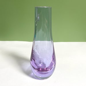 May include: A clear glass vase with a tapered neck and a rounded base, featuring a subtle purple tint at the bottom. The glass is decorated with an etched floral pattern on the front and includes a small rectangular sticker near the top rim.