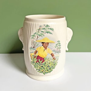 Può includere: Vaso in ceramica bianca con una scena di una persona con una camicia gialla e un cappello conico che raccoglie foglie di tè in un campo. Il vaso ha due manici a forma di elefante.