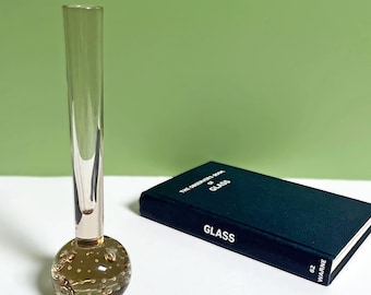 May include: A clear glass bud vase with a round, bubble-filled base and a slender neck. Next to it is a dark green book titled "GLASS". The vase is approximately 7 inches tall.
