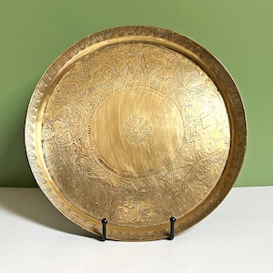 May include: A round brass tray featuring an intricate engraved pattern of stylized animals and floral motifs surrounding a central flower design. The rim is decorated with a repeating leaf pattern, and the metal surface has a polished gold tone.