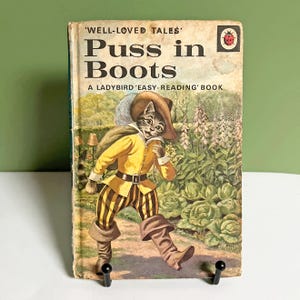 May include: Vintage Ladybird book titled "Puss in Boots." The cover features an illustration of a cat in a yellow jacket, striped pants, and boots, walking through a garden. The book has a worn appearance, with the title and author's name visible.