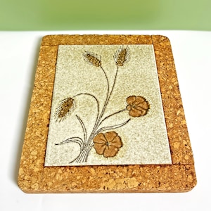 May include: A square cork trivet with a central ceramic tile. The tile features a brown floral design with wheat stalks and flowers on a speckled beige background. The cork border provides a natural, rustic aesthetic.