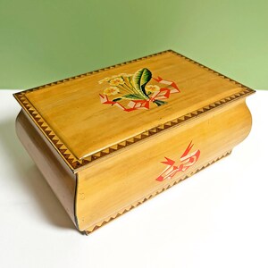 Könnte beinhalten: Eine hellbraune Holzbox mit floralem Design auf dem Deckel. Der Deckel hat eine dekorative Bordüre und ein Design aus gelben Blumen, grünen Blättern und roten Bändern. Die Box hat eine geschwungene Form und ein dreieckiges Muster an den Rändern.