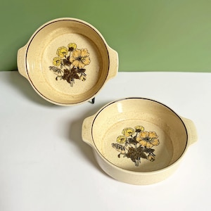May include: Two cream-coloured ceramic bowls with handles. Each bowl features a floral design with yellow and brown flowers. The bowls have a speckled texture and a brown rim, ideal for serving dishes.
