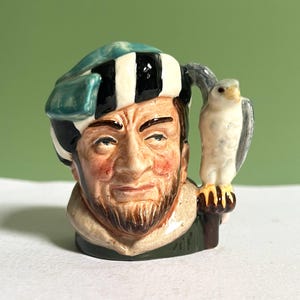 Peut inclure: Un pichet en céramique en forme de personnage portant une coiffe rayée bleu sarcelle, noire et blanche. La figurine a une barbe brune et un faucon blanc perché sur l'anse. La base est vert foncé avec un col beige clair autour du cou.