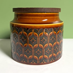 May include: A cylindrical ceramic storage jar featuring a dark brown and amber glaze with a repeating geometric pattern of interlocking shapes and central diamond motifs. The jar is topped with a flat, circular dark brown wooden lid.